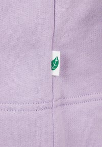 Material din bumbac de culoare mov deschis, cu un tricot texturat, având un eticheta albă cu o siluetă de frunză verde la cusătură.