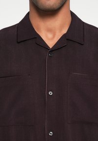 Sötét szilvaszínű rövid ujjú ing, gallérral, két elülső zsebbel és kontrasztos varrással díszített fekete gombokkal.