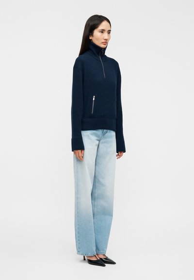 Polo à fermeture éclair marine en tissu doux, avec un col haut, des poches latérales zippées, associé à un jean en denim bleu clair à jambes larges.