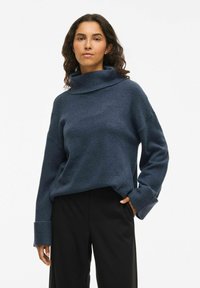 Pull à col roulé bleu marine en tissu texturé, avec des poignets larges et une coupe décontractée, associé à un pantalon noir.