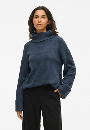 Pull à col roulé bleu marine en tissu texturé, avec des poignets larges et une coupe décontract�ée, associé à un pantalon noir.