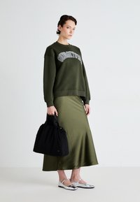Dunkelgrüner Pullover mit weißem Text, langer olivgrüner Rock, silberne Ballerinas und eine schwarze Tasche mit gerafftem oberen Teil. Glatte Texturen, lässiges Design.