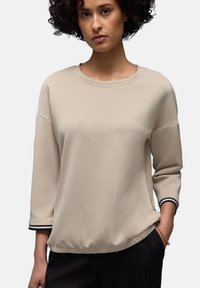 Frau mit lockigem Haar, die ein beiges Langarmshirt mit schwarz-weiß gestreiften Bündchen und eine dunkle Hose trägt, steht mit einer Hand in der Tasche.