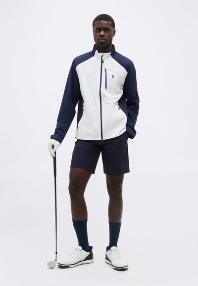 Hombre de pie sosteniendo un palo de golf, vistiendo chaqueta azul marino y blanca, pantalones cortos azul marino, calcetas azules marinas, zapatos de golf blancos y un guante blanco en la mano izquierda.