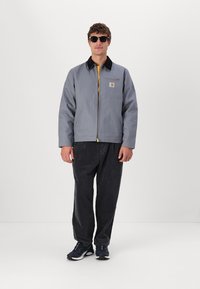 Veste zippée grise avec un intérieur jaune et un col noir, associée à un pantalon en velours côtelé sombre et des baskets bleu marine. Comprend un patch logo sur la poitrine.
