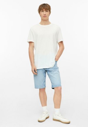 Junger Mann steht und trägt ein schlichtes weißes T-Shirt, hellblaue Jeansshorts, weiße Socken und weiße Freizeitsneaker vor weißem Hintergrund.