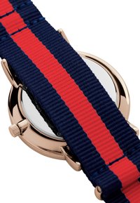 Marineblå stoffurrem med røde striper, med en rund roségullkasse og en reflekterende glassoverflate. Justerbar metallkrone synlig.
