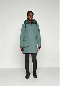 Vattentät teal parka med frontdragkedja, justerbar huva, två sidofickor och elastiska ärmslut; kombinerad med svarta byxor och skor.