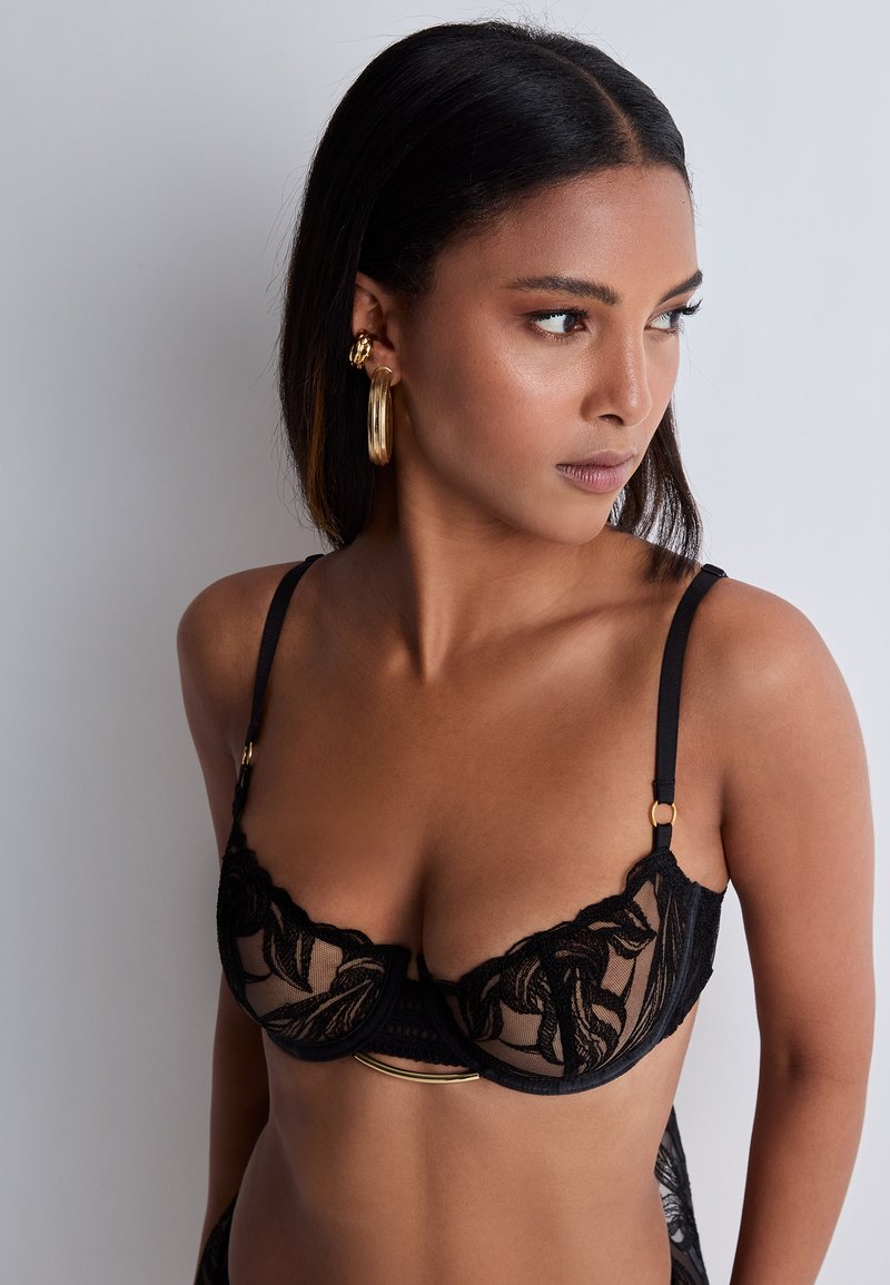 Soutien-gorge en dentelle noire avec un motif floral, des bonnets transparents, des accents en métal doré et des bretelles réglables pour un ajustement personnalisé.