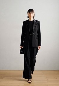 Černé sakové sako nošené přes černý top, spárované s volnými černými kalhotami a držící malé černé kabelku. Sandály dokončují tento outfit.