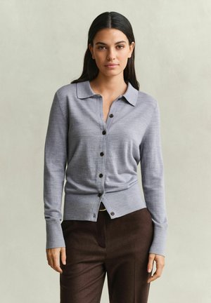 Femme aux longs cheveux foncés portant un cardigan gris clair boutonné et un pantalon marron foncé sur un fond uni.