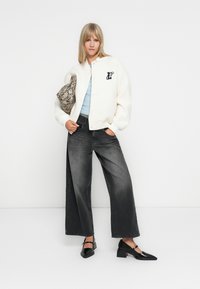 Veste bomber en mélange de laine blanche avec un col rond et un logo, haut ajusté bleu clair, jeans noir à jambes larges et ballerines noires à bout pointu. Pochette en cuir de serpent.