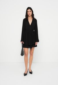 Μαύρο φόρεμα τύλιγμα με μακριά μανίκια, πλισέ υφή, ντεκολτέ σε σχήμα V και φουσκωτή κάτω πλευρά. Συνδυασμένο με μυτερά φλατ παπούτσια και τσάντα με πουά.