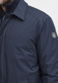 Chaqueta azul marino con cuello acanalado, cierre de cremallera y un parche con el logo en el hombro izquierdo. Fabricada con una tela lisa e insulating.