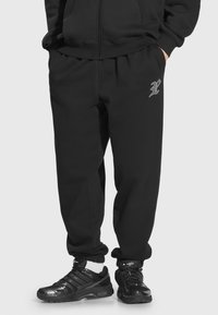 Czarne dresy z elastycznym pasem, zwężanymi kostkami i małym szarym logo na lewej udzie, w połączeniu z błyszczącymi czarnymi butami sportowymi.