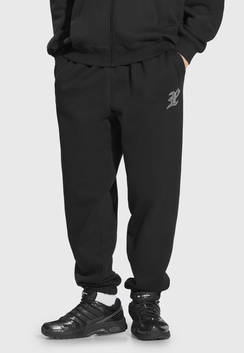 Czarne dresy z elastycznym pasem, zwężanymi kostkami i małym szarym logo na lewej udzie, w połączeniu z błyszczącymi czarnymi butami sportowymi.