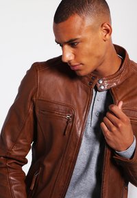 Chaqueta de cuero marrón con un cuello alto, bolsillos con cremallera y una textura suave. Lleva puesta una camiseta henley gris, que presenta un diseño ajustado.
