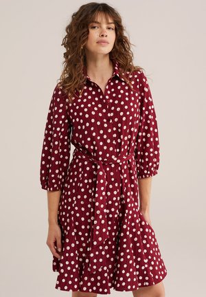 Femme portant une robe rouge à pois blancs, manches 3/4, devant boutonné, et une ceinture assortie en tissu à la taille.