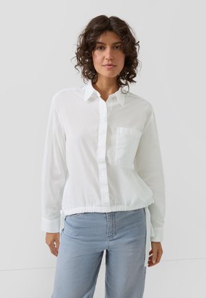 Kobieta z kręconymi brązowymi włosami, ubrana w białą koszulę na guziki z długim rękawem i kieszenią na piersi oraz jasnoniebieskie dżinsy, stojąca na jednolitym tle.