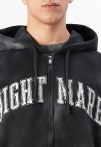 Close-up of a person wearing a black zip-up hoodie with white distressed text "NIGHT MARRE" and black drawstrings.