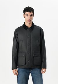 Veste noire en matériau lisse, avec un col, une fermeture à boutons et deux poches avant. Accents texturés sur le col.
