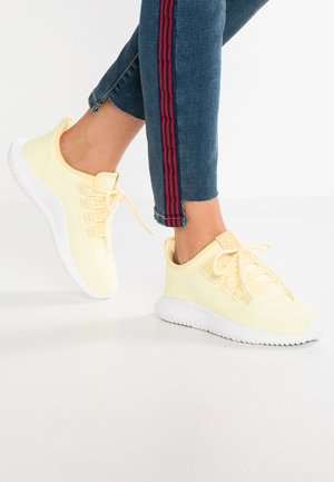 Personne portant des baskets tricotées jaune pâle avec des semelles blanches et un jean bleu avec des rayures rouges près des ourlets à la cheville.