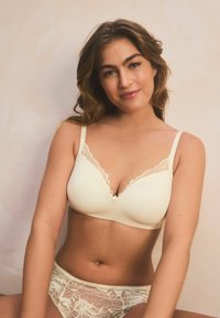 Soutien-gorge crème avec finition en dentelle, tissu lisse, bonnets rembourrés et bretelles réglables ; assorti à un ensemble de sous-vêtements en dentelle avec un motif floral.