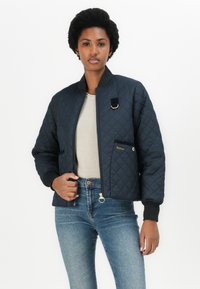 Navy quilted bomber jacket with black ribbed cuffs and collar, gold hardware accents, and a front pocket featuring the brand name "Barbour."