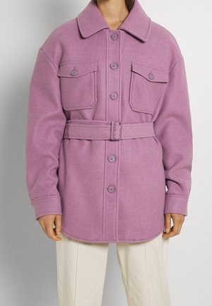 Kvinde iført en mauve uldfrakke med bælte, stor krave, knapper foran og to brystlommer med klap, kombineret med off-white bukser.