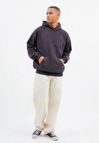 Welurowa bluza z kapturem w ciemnoszarym kolorze, z kieszenią kangurką, zestawiona z kremowymi spodniami o szerokich nogawkach i czarnymi trampkami.