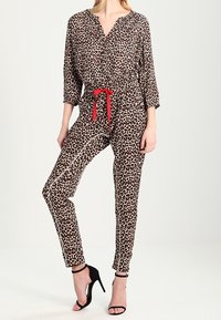Løveprint jumpsuit i lysebrun med sorte pletter; har lange ærmer, v-halsudskæring, rød bælte, og tætsiddende bukser. Sorte hæle er synlige.