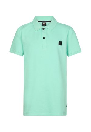 Jongen met krullend haar, die een muntgroen poloshirt, donkergrijze jeans en witte sneakers draagt, staat en kijkt opzij.