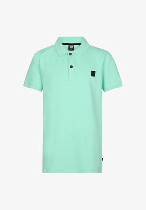 Jongen met krullend haar, die een muntgroen poloshirt, donkergrijze jeans en witte sneakers draagt, staat en kijkt opzij.