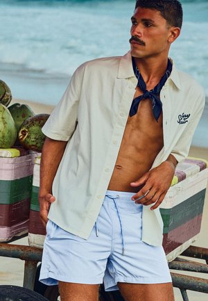 Bărbat purtând o cămașă bej deschisă, eșarfă neagră la gât și pantaloni scurți bleu deschis, sprijinindu-se de un cărucior cu nuci de cocos pe plajă.