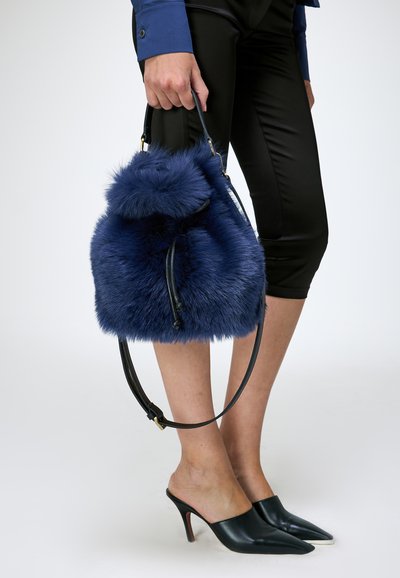 Personne tenant un sac à main bleu duveteux, portant un pantalon noir court, une chemise bleue à manches longues et des mules noires à talons sur fond blanc.