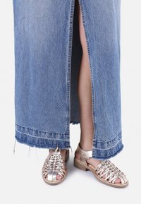 Falda maxi de denim con dobladillo deshilachado y abertura lateral. Sandalias de plata trenzadas con tacón de madera que muestran un diseño texturizado y abierto.