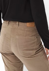 Beige Corduroy-Hosen mit glatter Textur, ausgestattet mit Gesäßtaschen und einem braunen Lederetikett mit der Aufschrift "Raphaela by Brax."
