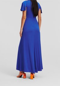 Kvinna klädd i en lång klänning i kunglig blå färg med korta fladdrande ärmar och orange högklackade sandaler, stående med ryggen mot kameran på en neutral bakgrund.