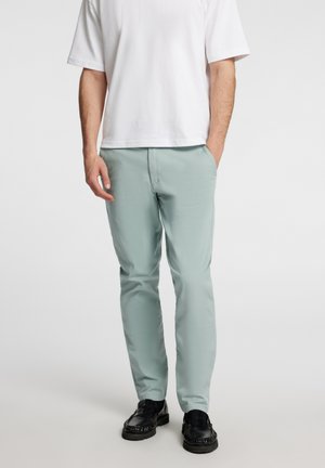 Pantalons vert clair en coupe slim avec une texture lisse, dotés de poches latérales et d'un design épuré, associés à des chaussures noires et une chemise blanche.