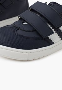 Tumedate sinised tennisetossud valgete aktsentidega, valmistatud tekstuuriga materjalist, millel on kaks Velcro-riba ja kummist tald. Üksikasjalik vaade.