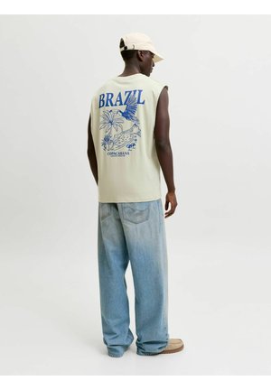 Homme portant une chemise beige sans manches avec un imprimé bleu Copacabana Brésil, un jean ample bleu clair, des chaussures beiges et une casquette beige, de dos.