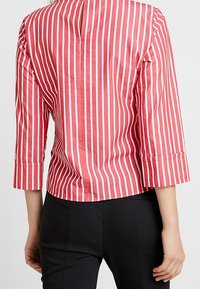 Camicia a righe rossa e bianca con maniche a tre quarti, una piccola apertura sul retro e texture liscia. Indossata con pantaloni neri.