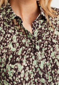 Kragarskjorta med ett grönt och svart prickigt mönster. Tillverkad av ett lätt tyg med en mjuk textur och knappstängning.