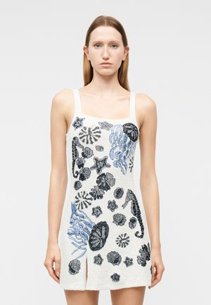 Mujer vestida con un vestido blanco sin mangas con bordados en azul y negro de criaturas marinas y conchas, posando frente a un fondo liso.