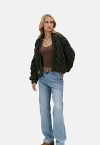 Gruby zielony kardigan z grubym splotem warkoczowym i oversize'owymi rękawami, zestawiony z brązowym topem na ramiączkach i jasnoniebieskimi szerokimi jeansami.