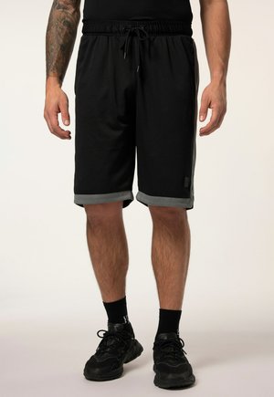 Mand iført sorte netshorts med grå manchetter, sorte sokker og sorte sneakers stående mod en ensfarvet lys baggrund.