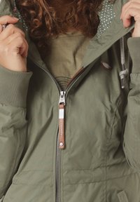 Giacca puffer verde oliva con chiusura a zip, polsini a coste, cappuccio con lacci e fodera a pois. Presenta dettagli in pelle marrone.