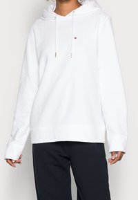 Biały hoodie wykonany z miękkiego materiału, z kieszenią z przodu, regulowanym kapturem z metalowymi końcówkami oraz ściągaczami przy mankietach. Minimalistyczny detal logo na klatce piersiowej.