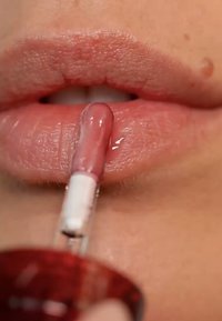 Gloss à lèvres dans un tube transparent avec un applicateur blanc, appliqué sur des lèvres douces de couleur nude, mettant en valeur une finition brillante et une texture lisse.