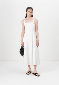 Robe midi en lin blanc avec décolleté carré et fines bretelles, associée à des tongs noires et un petit sac pochette noir. Design simple et épuré.